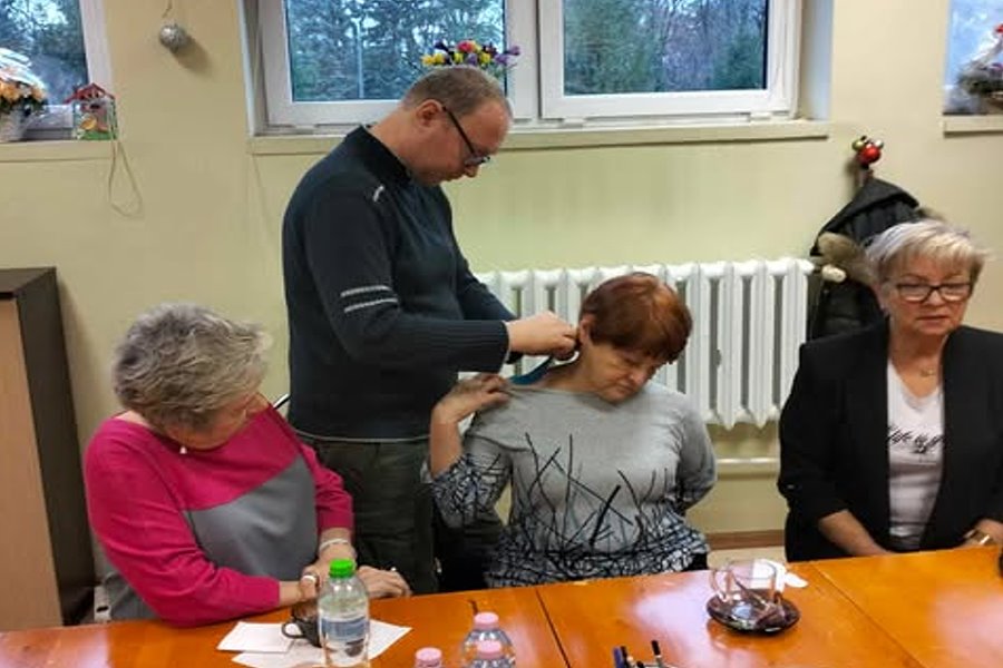 Mężczyzna nakłada taśmę rehabilitacyjną na szyję kobiety siedzącej przy stole; wok&oacute;ł niego kilka starszych os&oacute;b uczestniczących w zajęciach w sali.