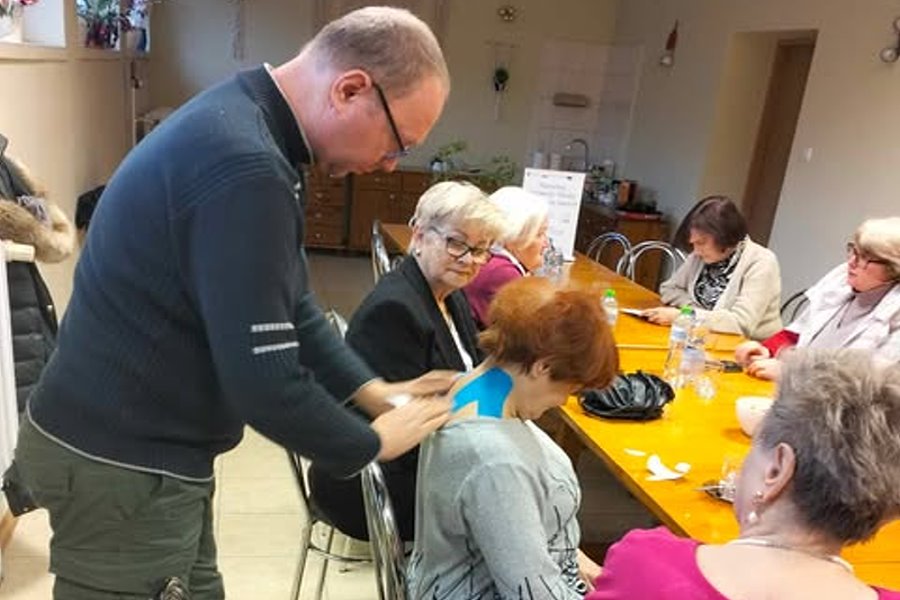 Mężczyzna nakłada taśmę rehabilitacyjną na szyję kobiety siedzącej przy stole; wok&oacute;ł niego kilka starszych os&oacute;b uczestniczących w zajęciach w sali.